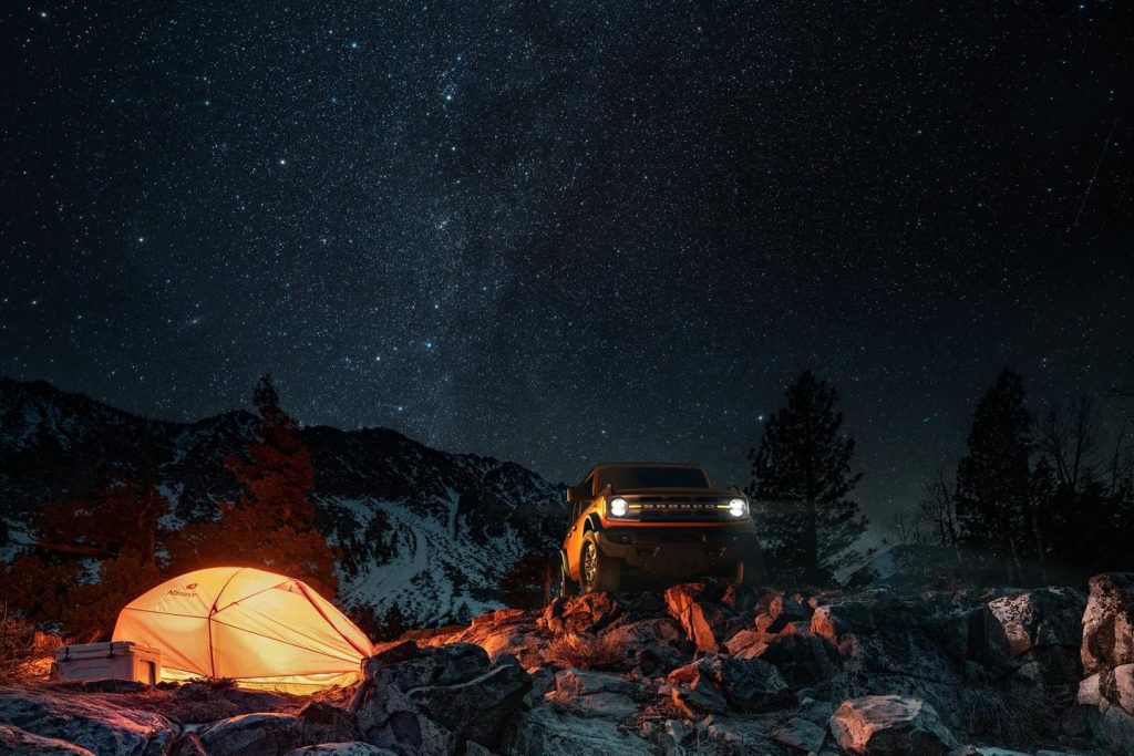 A formidable image of the starry, night sky, with the 2022 Ford Bronco blaring its headlights in the dark. The image of a lit tent and snowy mountains can be seen.
