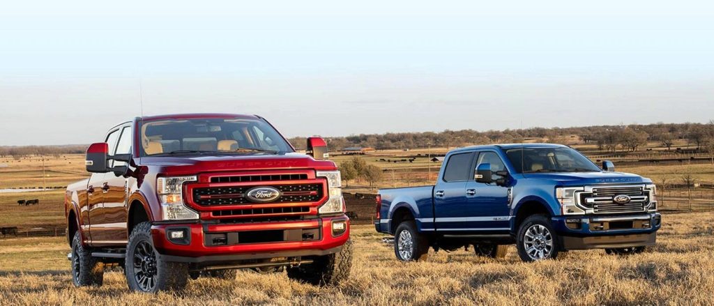 A photo of two 2022 Ford F-250 trucks, Red on the left, and a blue on the right, parked on a field with yellow grass.