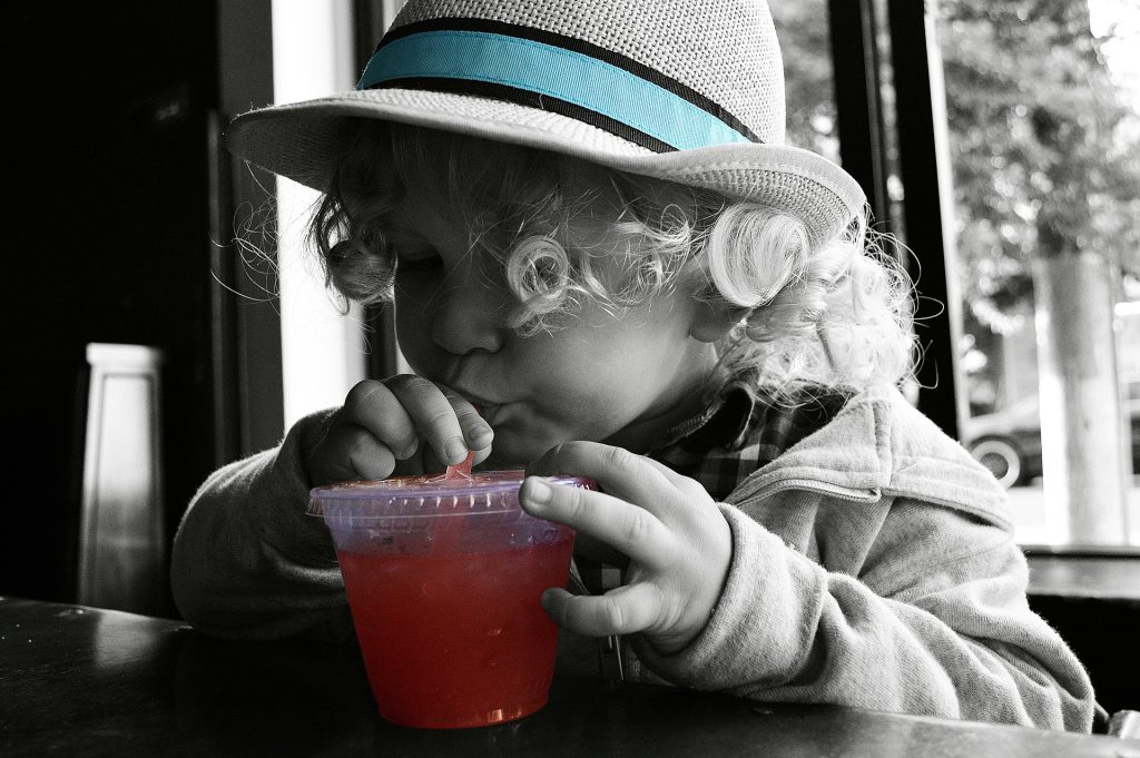 A black and white photo of a child with colors only being emphasized on the blue strip of their hat and the red color of their drink.