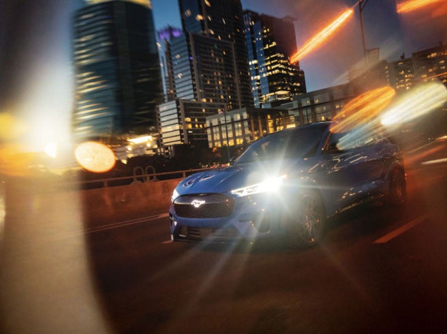 Blue 2024 Ford Mustang Mach-E driving with a cityscape behind it. 